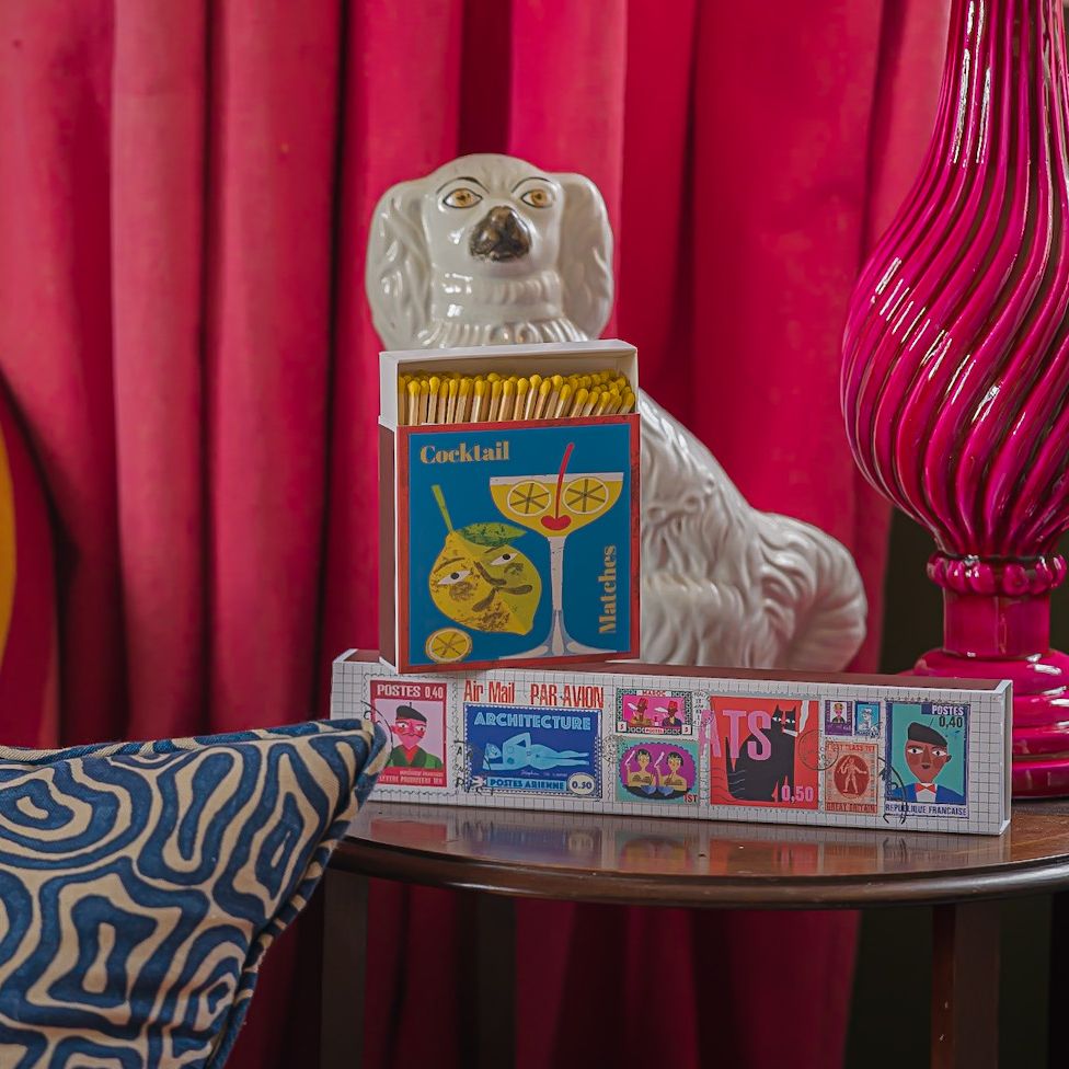 Cocktail matchbox stacked on a box of long matches in front of a china dog on a side table