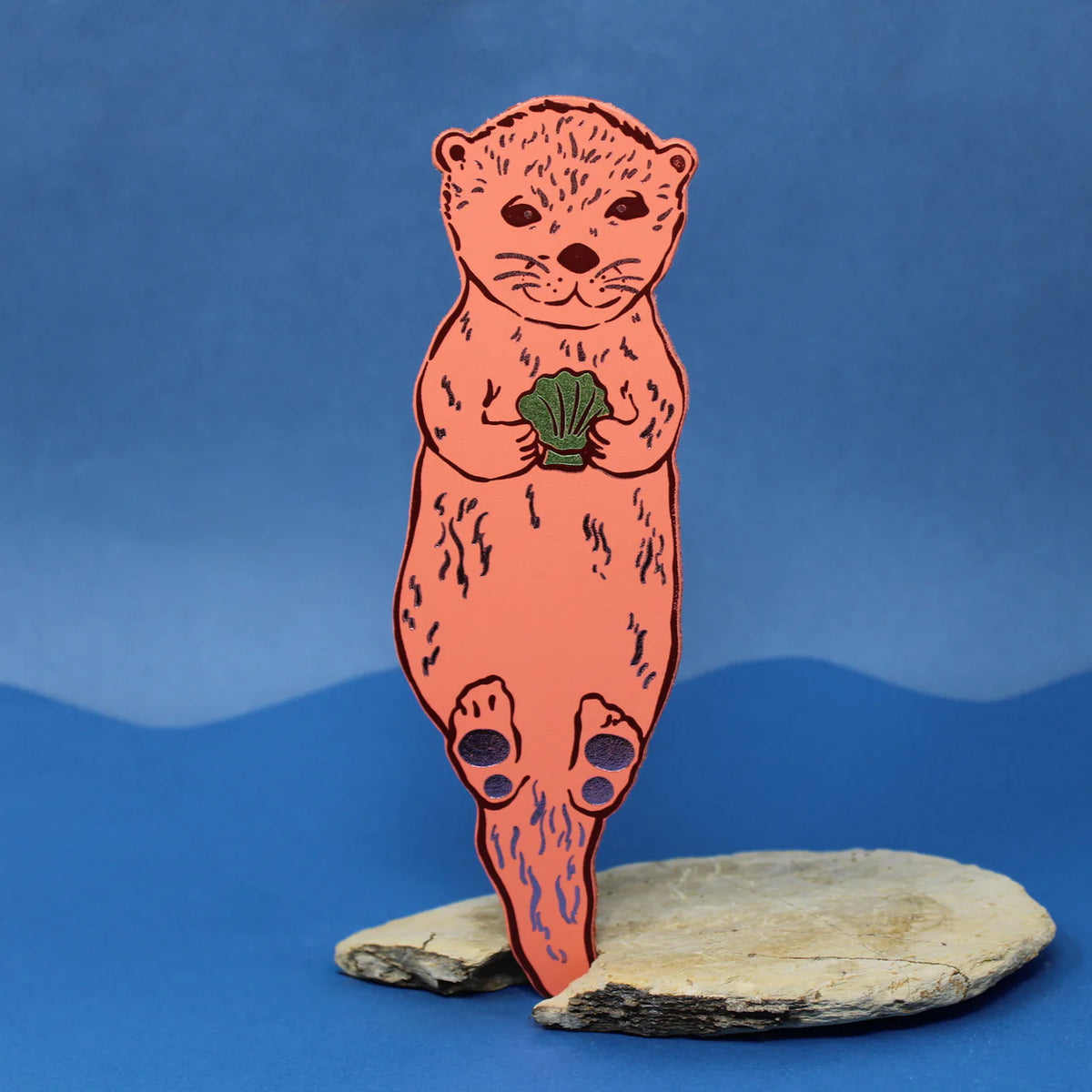 Ark apricot otter bookmark holding a shell, standing on a rock against blue