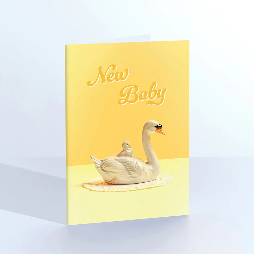 Ornament card: a vintage swan figurine with a cygnet on its back, with 'New baby' on yellow