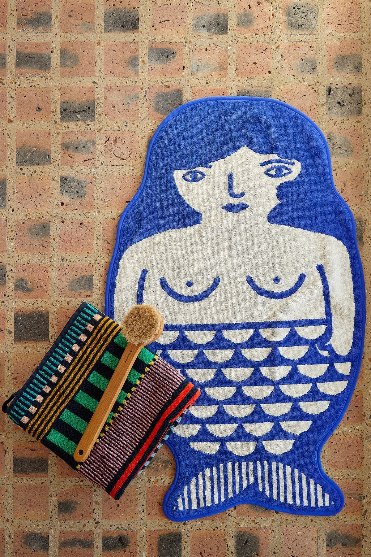 Mermaid bath mat on a brick floor with a striped towel and brush