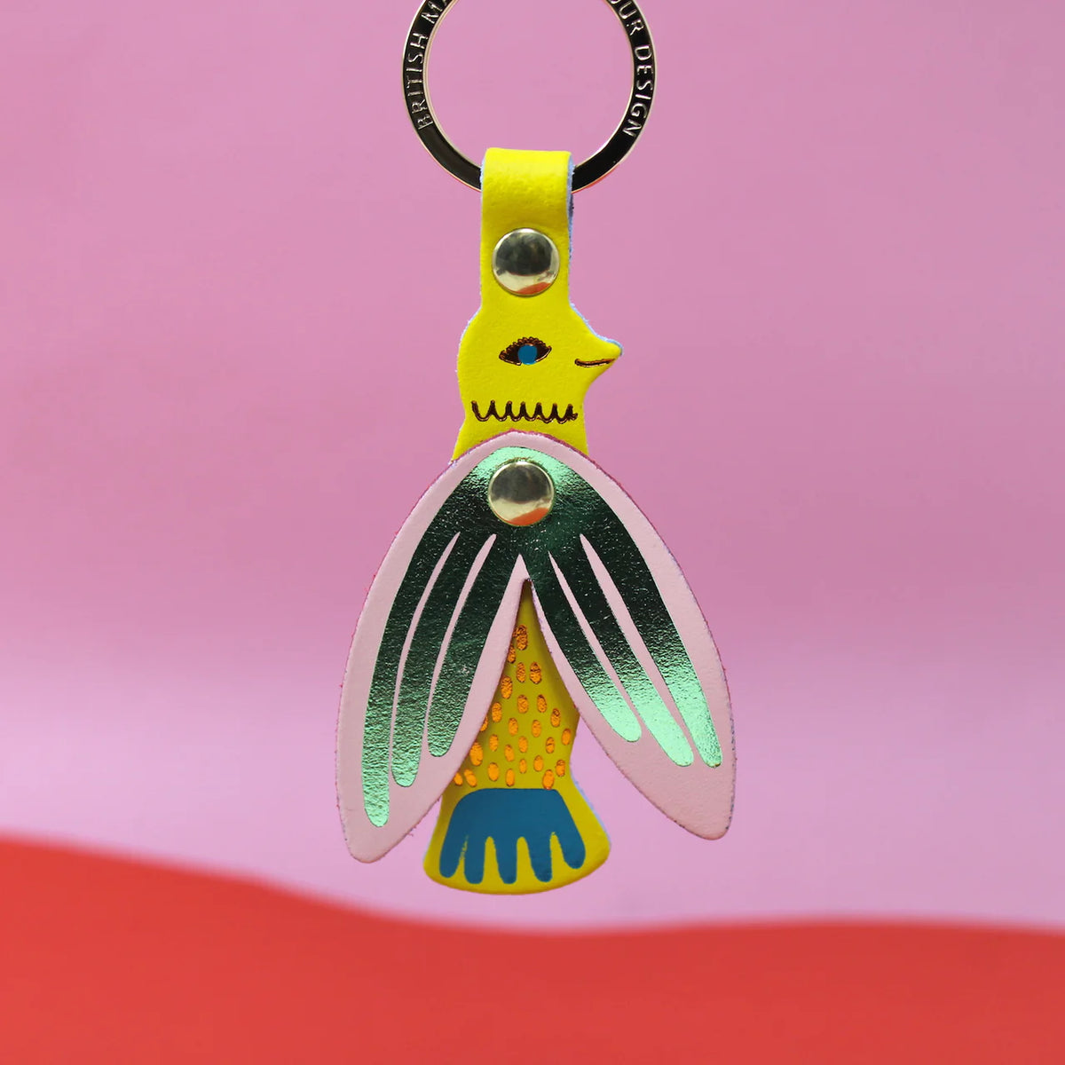 Yellow leather songbird key fob with green and pink metallic wings and a blue tail, on a black key ring, against a pink background