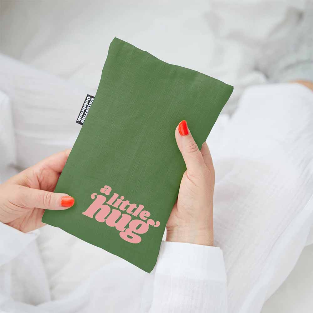 Hands holding a green linen hot water bottle cover with 'A little hug' in pink