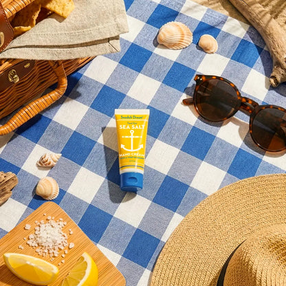 Swedish Dream hand cream on a blue gingham picnic blanket with shells, lemon and sunglasses