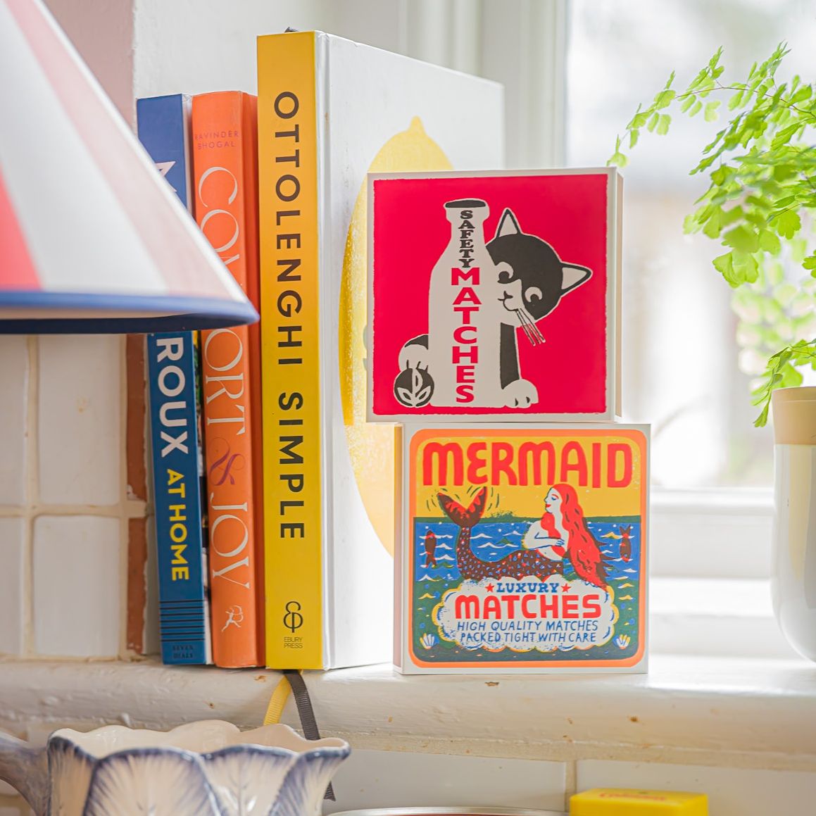 Mermaid matches stacked under a pink cat matchbox on a bookshelf beside cookbooks