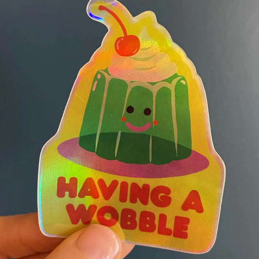Naomi Wilkinson holographic sticker: a smiling green jelly with a cherry, with 'Having a wobble'