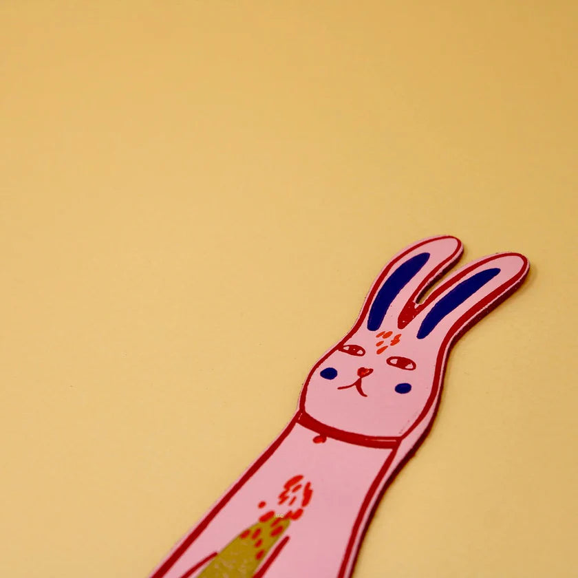 Close-up of an Ark bunny bookmark: a pale pink rabbit with navy ears and a red collar, on a mustard background