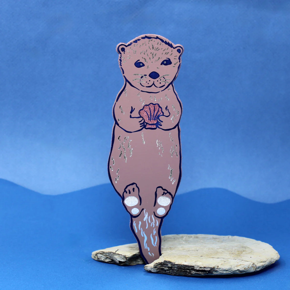 Ark dusky pink otter bookmark holding a shell, standing on a rock