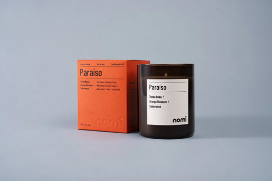Nomi Paraíso scented candle in an amber jar beside its orange box