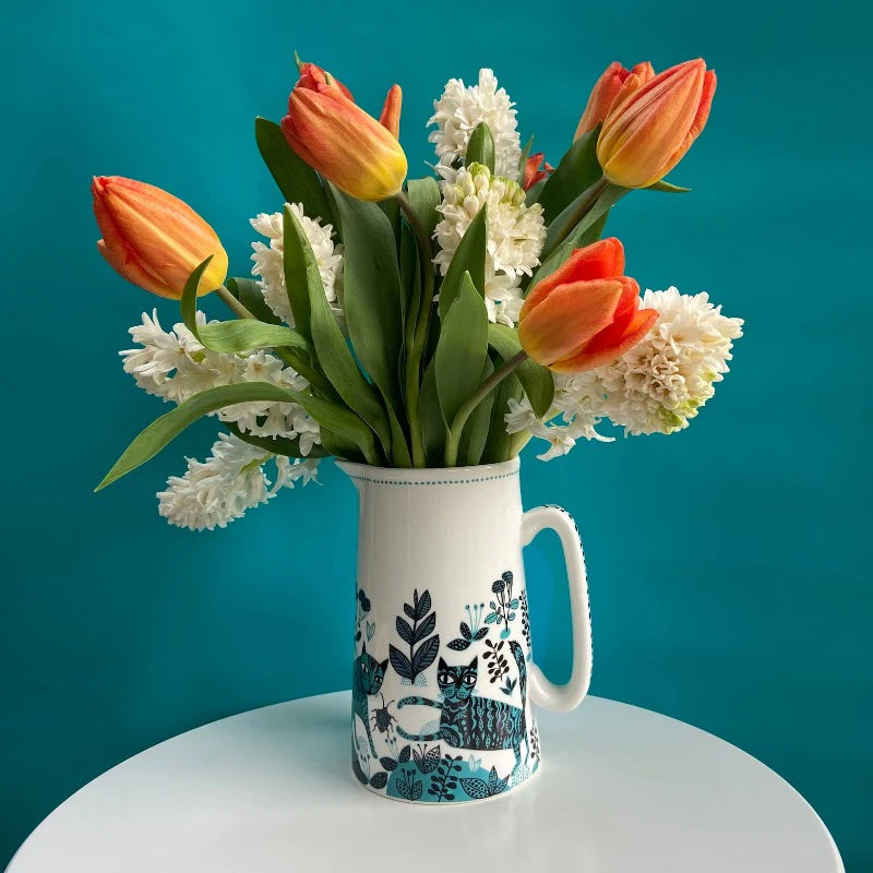 White ceramic jug painted with blue patterned cats, filled with orange tulips and hyacinths