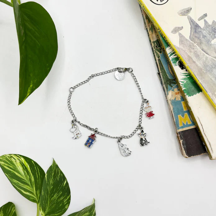 Silver chain bracelet with enamel Moomin family charms, laid flat beside a leaf
