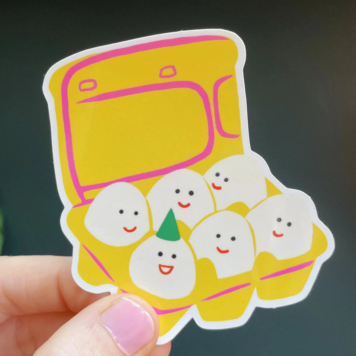 Naomi Wilkinson sticker of a yellow egg box with smiling eggs, one in a party hat, held in a hand