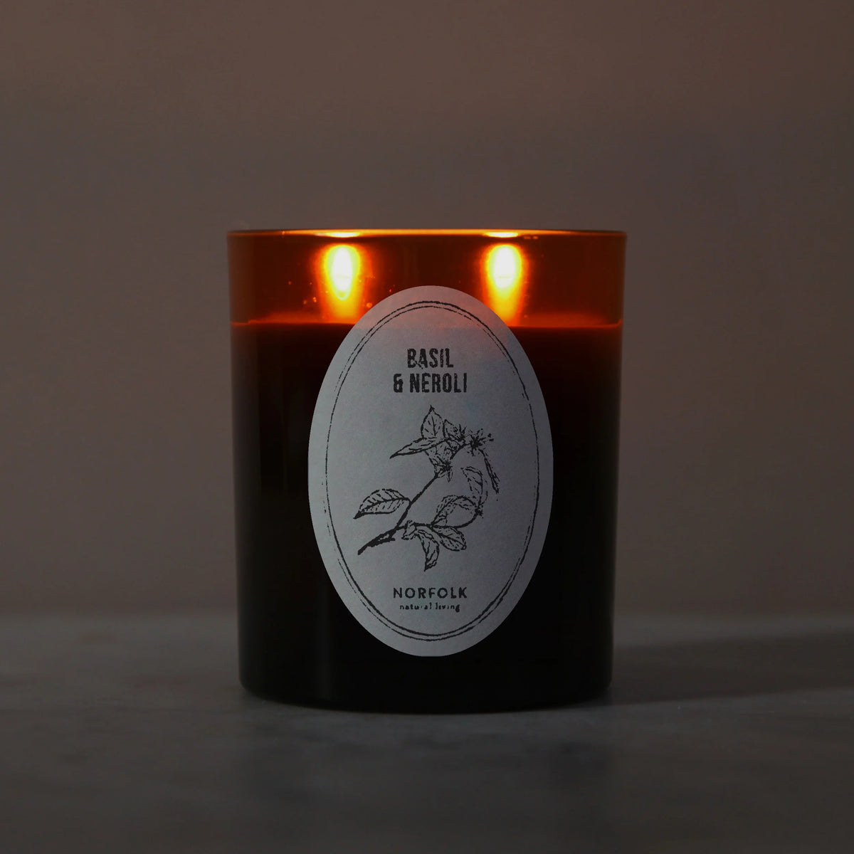 Basil & Neroli candle lit, with two flames glowing through the amber glass