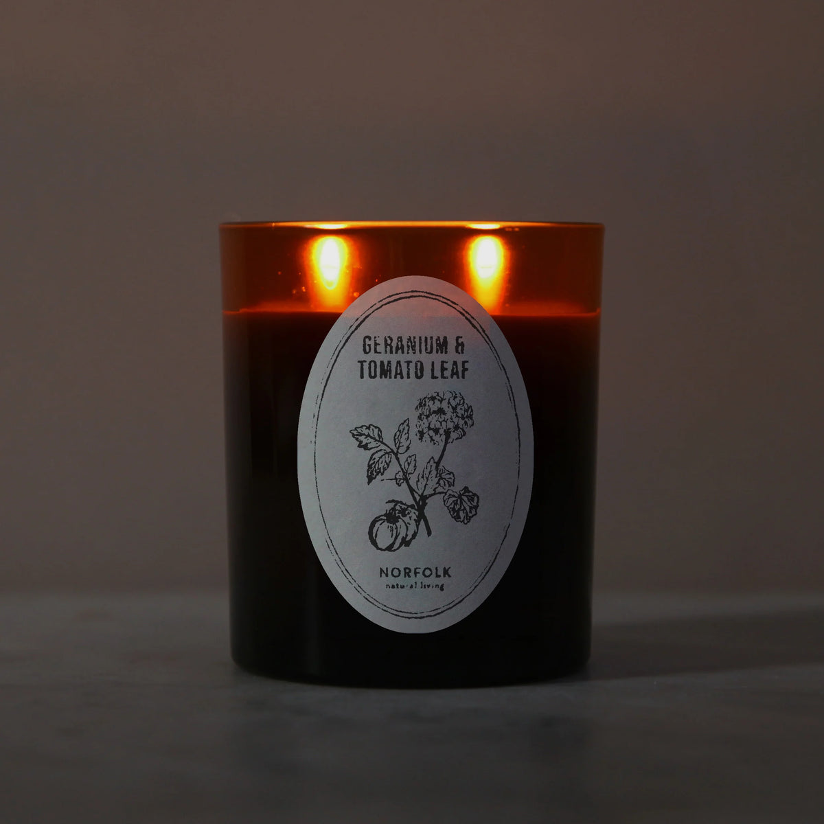 Geranium & Tomato Leaf candle lit, with two flames glowing through the amber glass