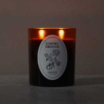 Geranium & Tomato Leaf candle lit, with two flames glowing through the amber glass