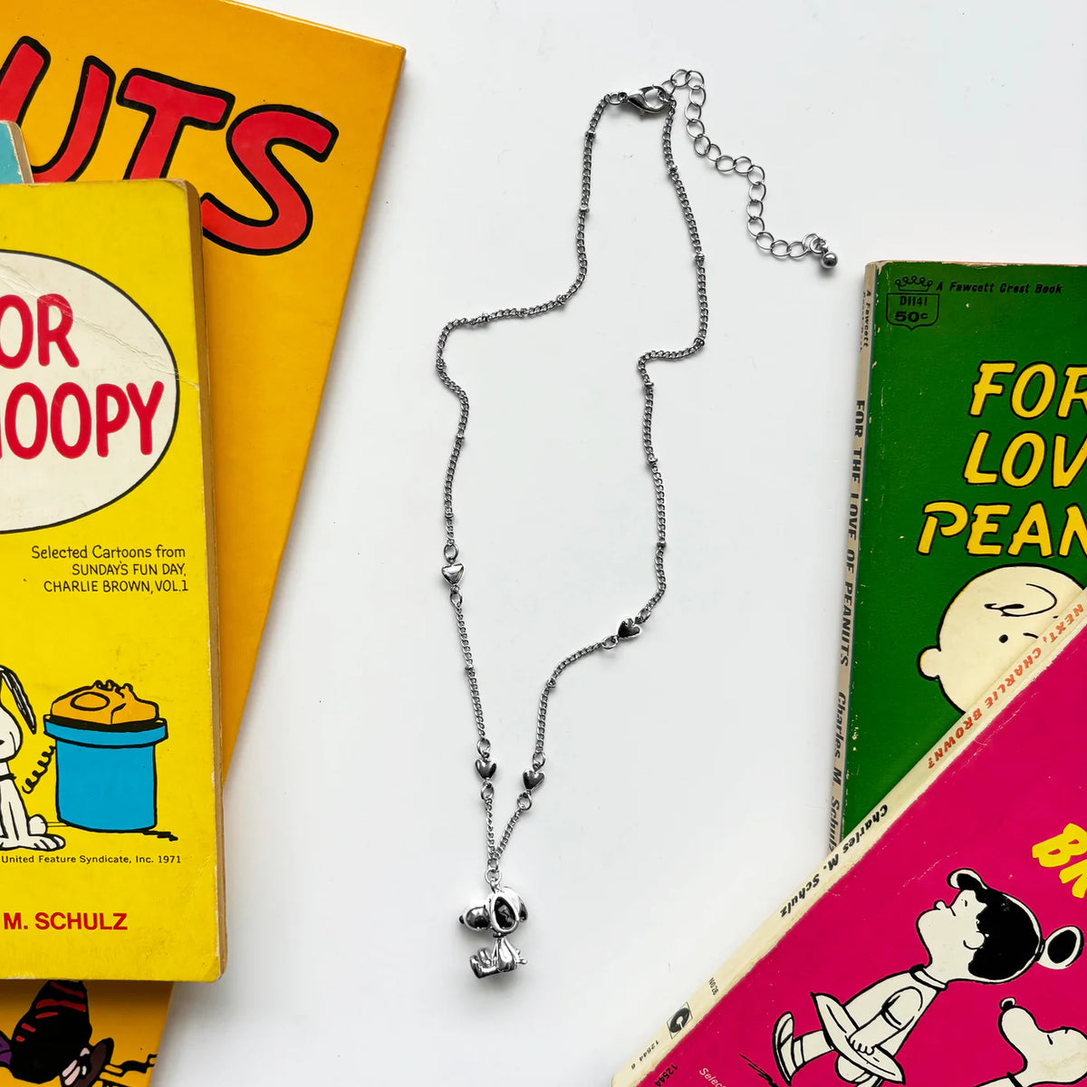 Silver chain necklace with a small 3D Snoopy charm, laid on Peanuts books