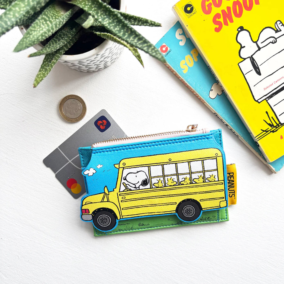 Peanuts card purse shaped like a yellow school bus with Snoopy and Woodstocks, beside a bank card and Snoopy book