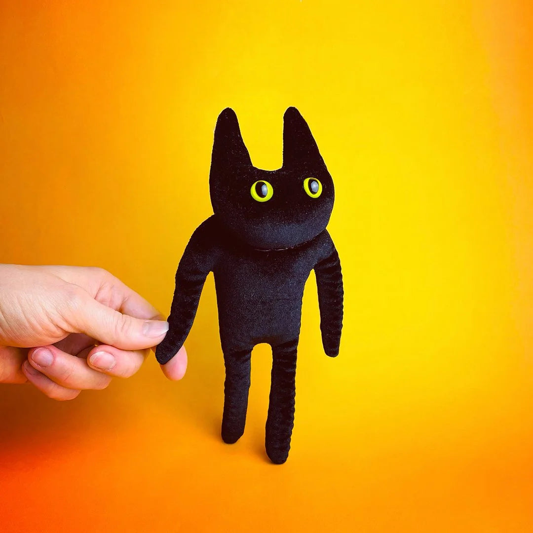 Mini Jasper by Ruby Ruth: a soft black cat toy with long limbs and yellow eyes, held up against orange