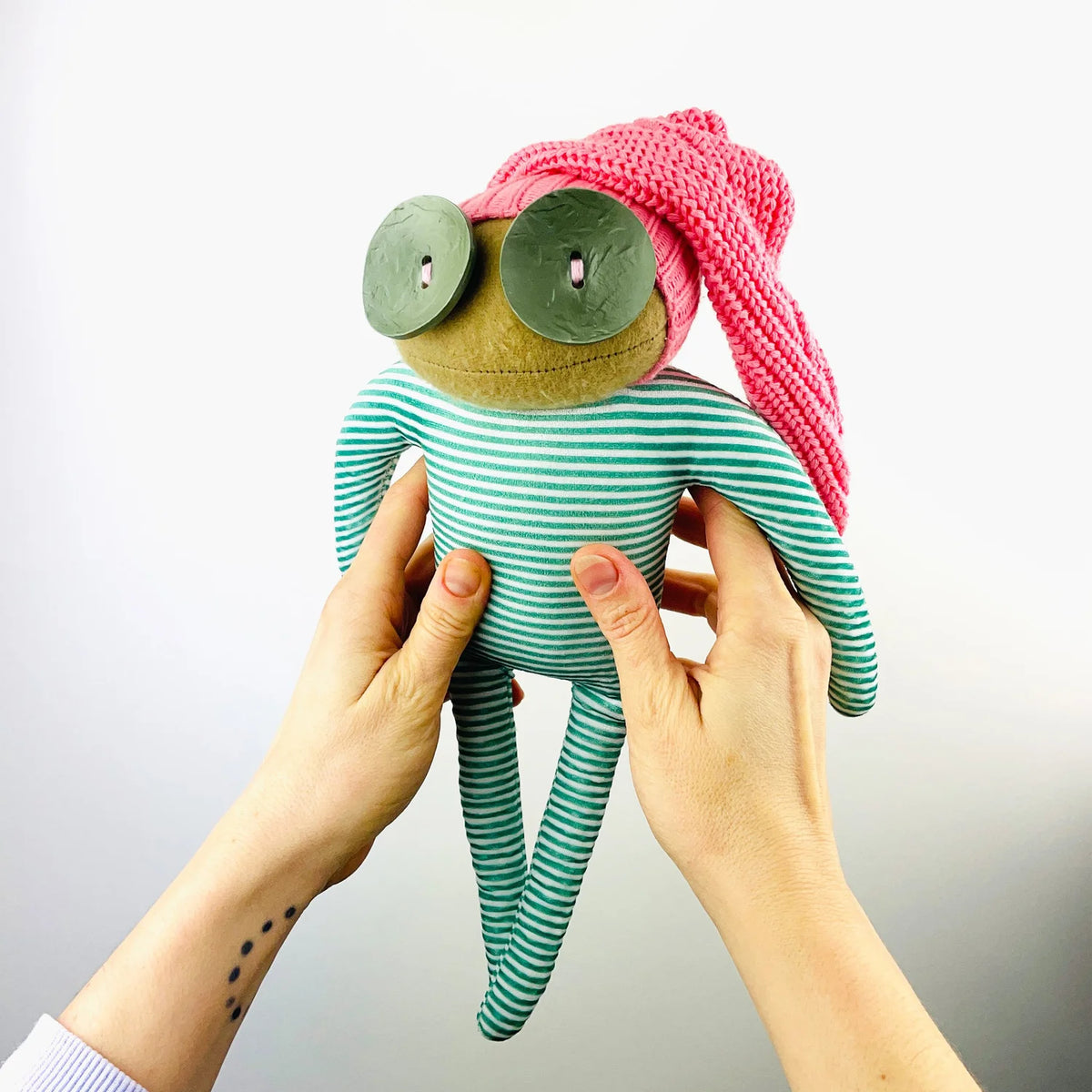 Ruby Ruth Rita soft toy: a creature with big green button eyes, a pink knitted hat and green striped limbs, held up