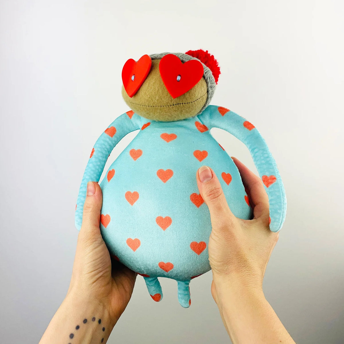 Light blue soft toy with red hearts, heart glasses and a red pompom hat, held in two hands