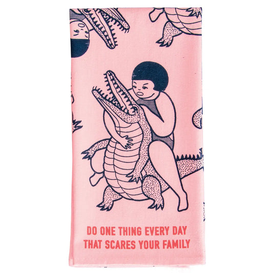 Blue Q tea towel, folded: a figure hugging a crocodile, with 'Do one thing every day that scares your family'