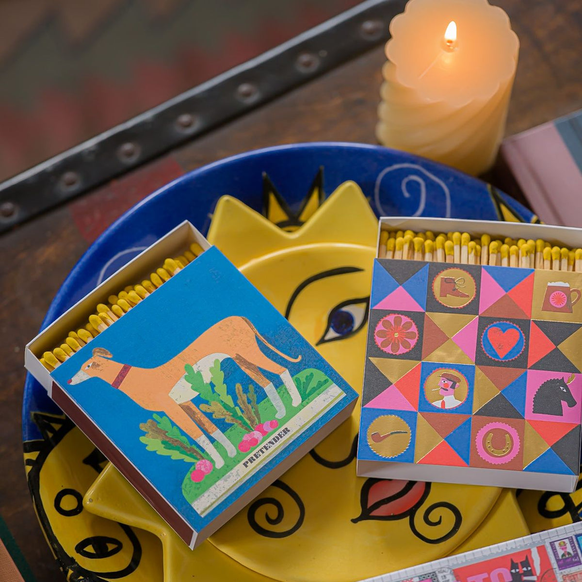 Whippet matchbox and a geometric patterned matchbox on a yellow sun plate beside a lit candle