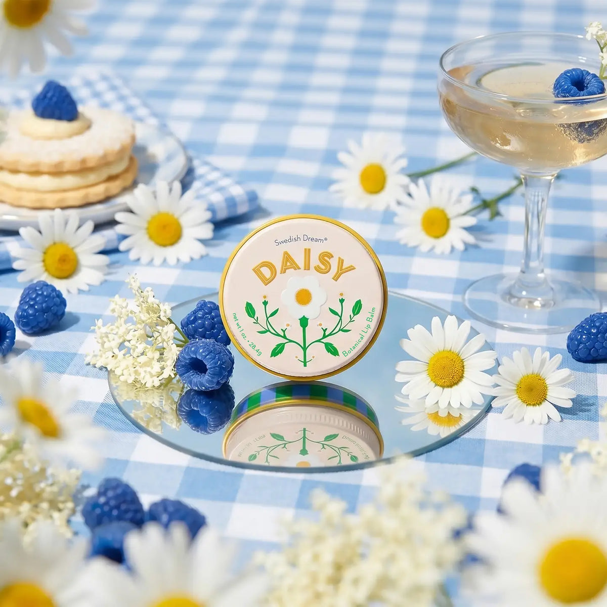 Daisy lip balm on a blue gingham picnic with daisies, berries and biscuits