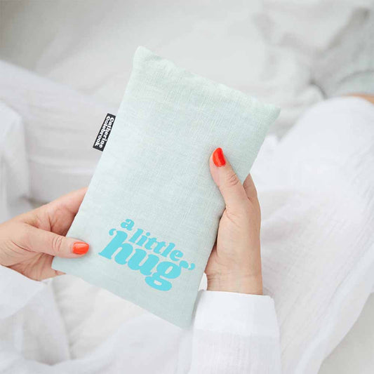 Hands holding a mint linen hot water bottle cover with 'A little hug' in blue