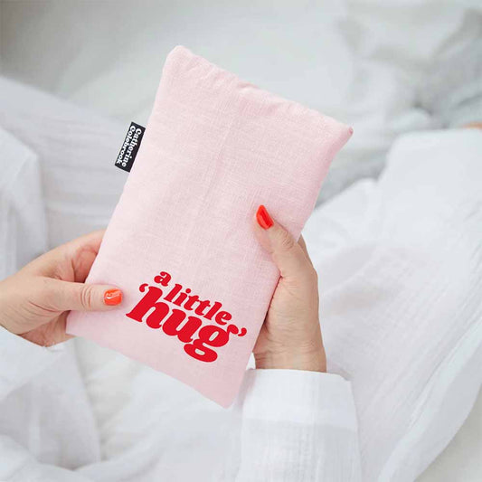 Hands holding a pink linen hot water bottle cover reading 'A little hug' in red