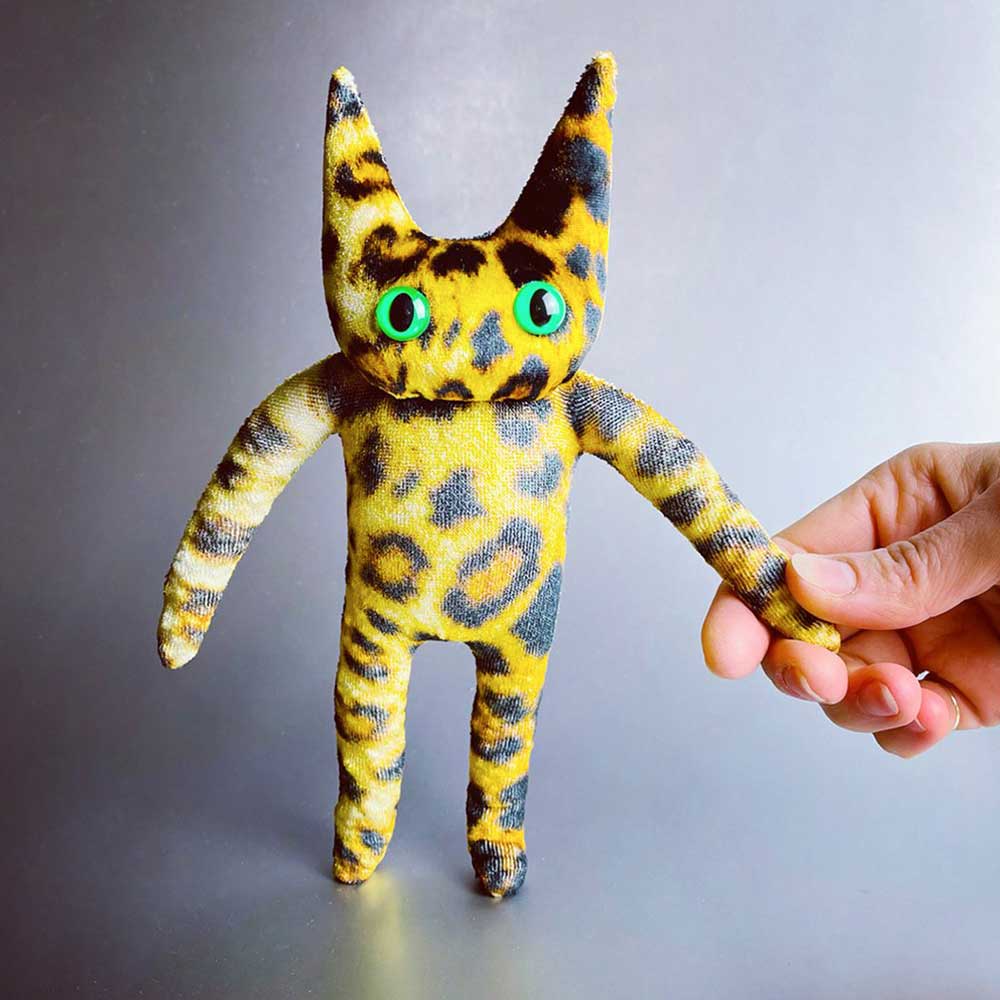 Small yellow and grey leopard print cat soft toy with green eyes, held by the arm