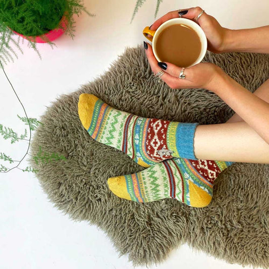 Person wearing yellow Fair Isle Moomin socks on a sheepskin rug, holding tea