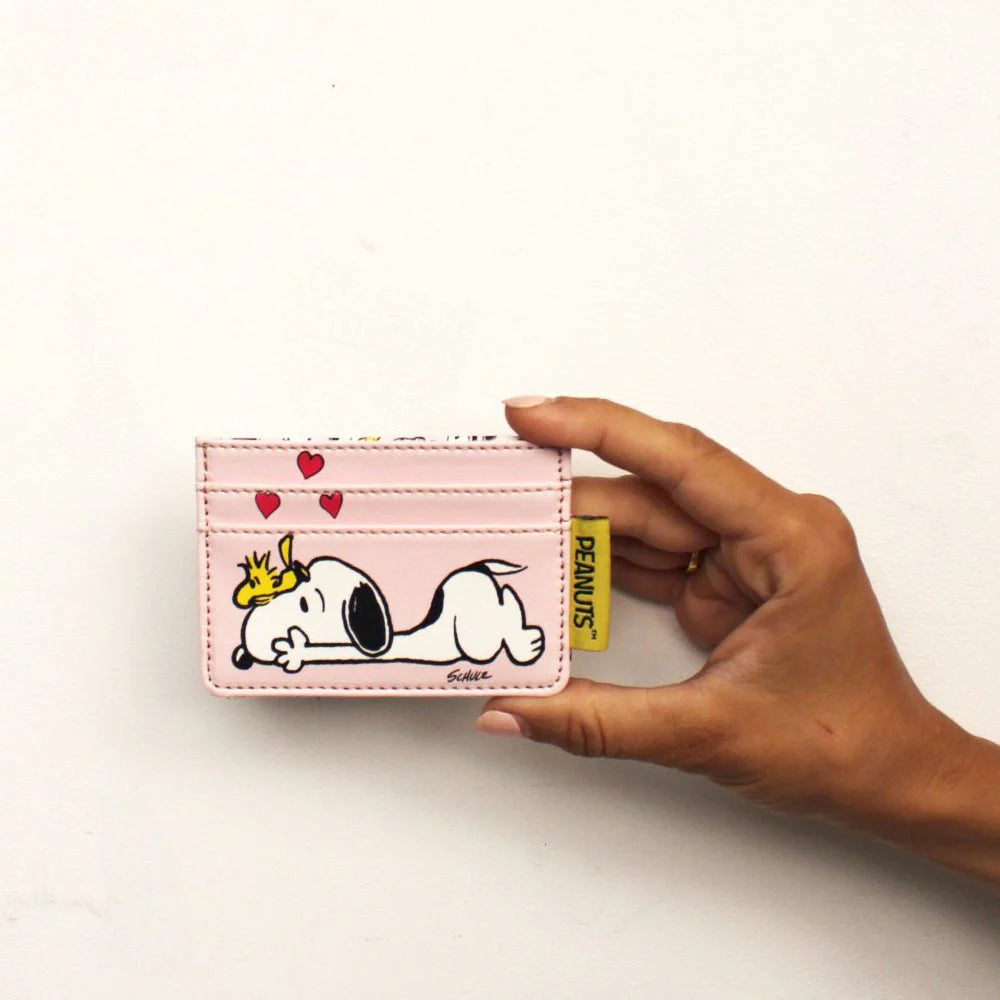 Hand holding a pale pink Peanuts card holder printed with Snoopy lying down, Woodstock on his head and red hearts above