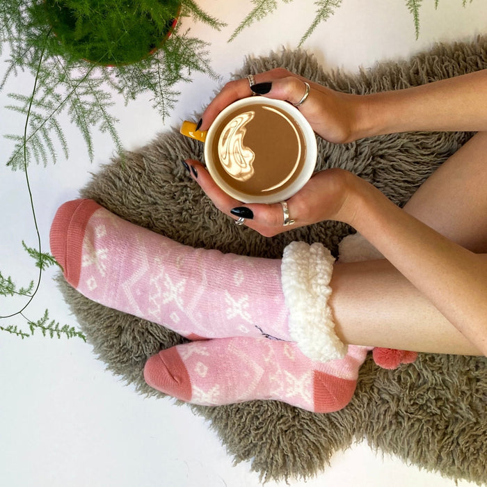Person in pink Moomin slipper socks holding a mug of coffee on a sheepskin rug