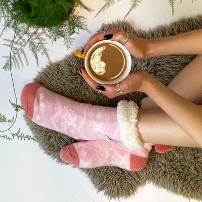 Person in pink Moomin slipper socks holding a mug of coffee on a sheepskin rug